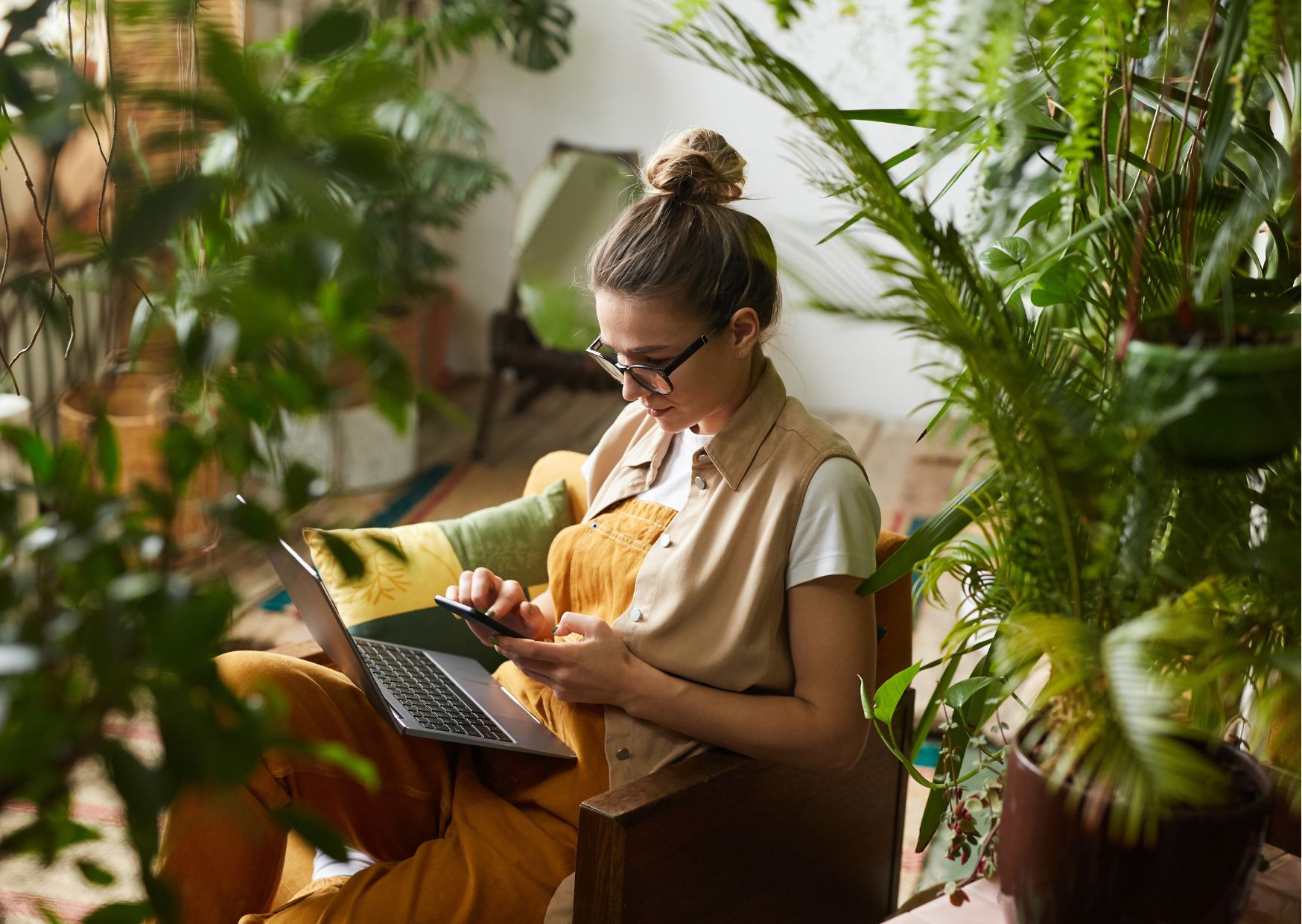 A women on her phone and computer around a bunch of houseplants.