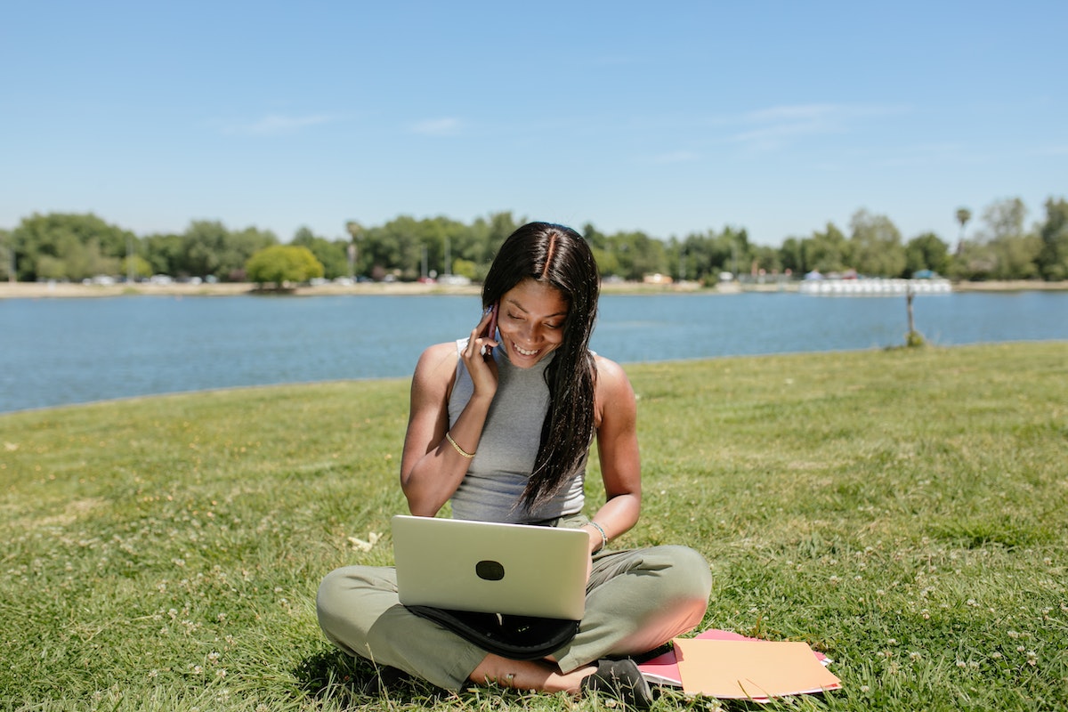 A woman sitting in the grass smiling while on her laptop and phone.